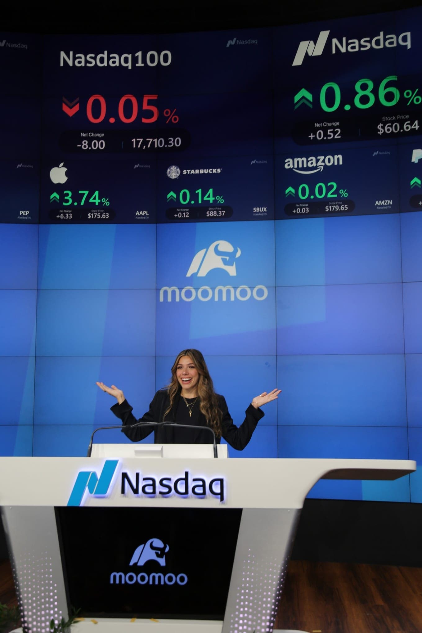 Taylor Price at the Nasdaq MarketSite podium for a Moomoo x Nasdaq appearance, with Moomoo and Nasdaq branding on the screens behind her.